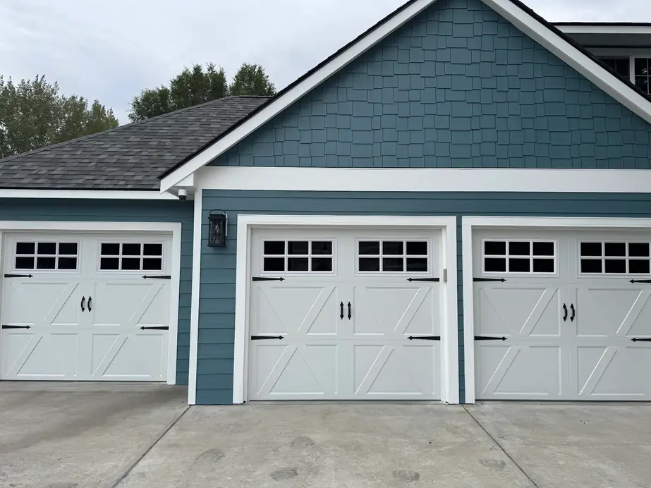 Three white carriage-house garage doors with X-bracing and grid windows on a slate-blue shingle house